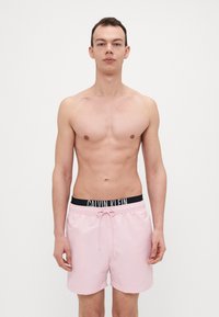 Helle rosa Badehosen aus glattem Stoff haben einen schwarzen Bund mit dem "CALVIN KLEIN" Logo und einen Kordelzug zur Anpassung.