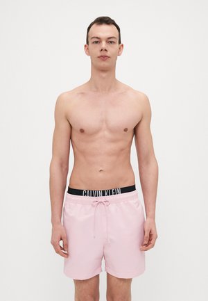 Lyse rosa badebukser laget av glatt stoff har et svart liv med "CALVIN KLEIN" logo og snor for justering.