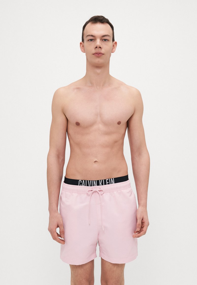 Helle rosa Badehosen aus glattem Stoff haben einen schwarzen Bund mit dem "CALVIN KLEIN" Logo und einen Kordelzug zur Anpassung.