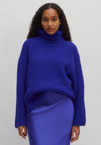 Oversized purple knit sweater with a high turtleneck and ribbed texture, layered over a silky blue skirt. Sweater has dropped shoulders.