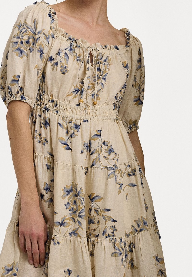 Blumenkleid aus Leinen mit Puffärmeln, quadratischem Ausschnitt und einer Kordelzugbindung. Beiger Grundton mit blauen und gelben Blumenmustern. Gestufter Design.