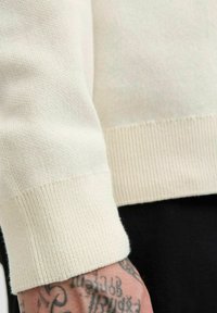 Helles cremfarbiges Sweatshirt mit strukturierter Strickmuster, ausgestattet mit gerippten Bündchen und Saum. Nahaufnahme eines Ärmels an einer tätowierten Hand.
