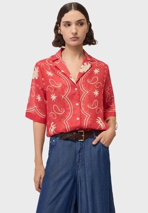 Kvinde med kort, bølget hår iført en rød mønstret skjorte med knapper, der er stoppet ned i højtaljede blå jeans med et brunt bælte.