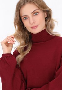 Burgunderfarbener Rollkragenpullover mit geripptem Muster und Puffärmeln. Das Model hat wellige blonde Haare und trägt kleine Creolen.