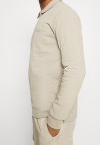 Chemise beige à manches longues avec un col zippé, une poche poitrine et des poignets côtelés. Tissu doux avec une coupe décontractée et confortable.