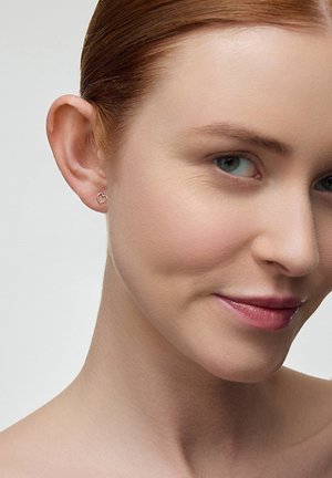 Złota, sercowa kolczyk wkręcany przymocowany do lewego ucha. Gładka powierzchnia, minimalistyczny design i niewielki rozmiar podkreślają subtelną elegancję.