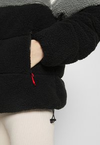 Pullover in pile nero e grigio con zip parziale frontale, dotato di tasca frontale con cursore rosso e orlo regolabile.