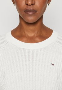 Maglione bianco lavorato a maglia con un motivo a trecce testurizzate e scollatura a girocollo a coste. Presenta un piccolo logo in basso a sinistra.