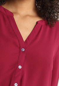Rode, lichte blouse met een V-hals en knopenlijst. Kenmerkt zich door kleine, lichte knopen en een zachte textuur.