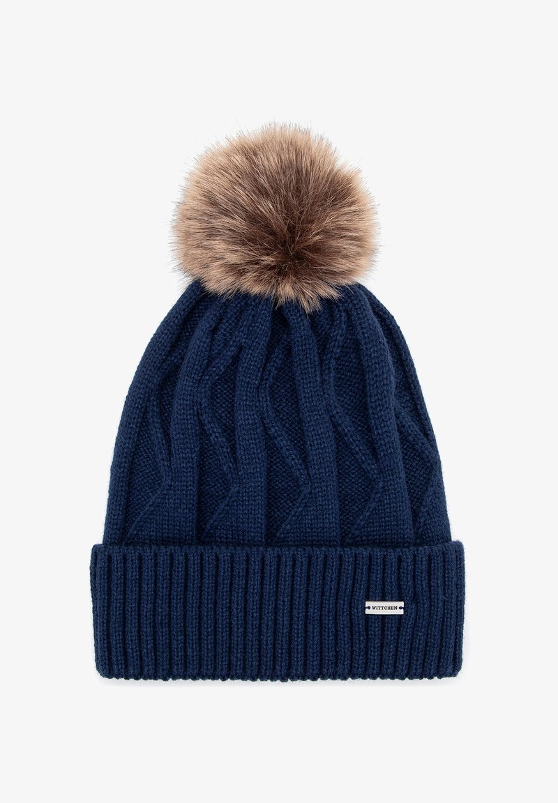 Námořnicky modrá pletená čepice s texturovaným vzorem copu a světle hnědým umělým kožíškem na vrcholu; na boku má malou značkovou cedulku.