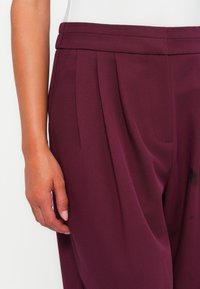 Pantaloni plissettati color bordeaux realizzati in tessuto morbido, con vita e tasche laterali, caratterizzati da una leggera lucentezza e linee pulite.