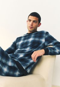 Bărbat tânăr cu păr scurt și închis la culoare, reclinându-se pe un scaun bej, purtând o cămașă cu mânecă lungă în carouri alb-albastru și pantaloni asortați, privind în lateral.