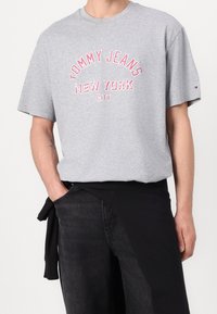 Szara bawełniana koszulka z krótkim rękawem z napisem "TOMMY JEANS NEW YORK CITY" w różowym kolorze. Okrągły dekolt, luźny krój, w połączeniu z czarnymi dżinsami.