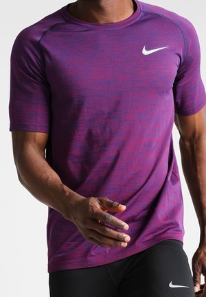 Torse masculin portant un t-shirt de sport à manches courtes violet et un short noir, tous deux arborant un logo Nike blanc.
