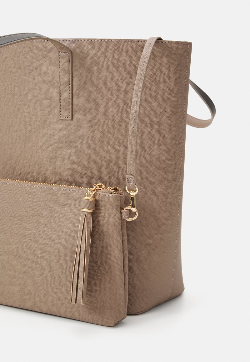 Bolso tote de color marrón claro con material texturizado, un bolsillo frontal con cremallera y un adorno de borlas doradas en el tirador de la cremallera. Incluye una correa de hombro ajustable.