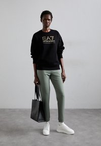 Svart sweatshirt med guld-logotyp, gröna tajta byxor, vita sneakers och en svart texturerad handväska. Slät textur med ett avslappnat design.