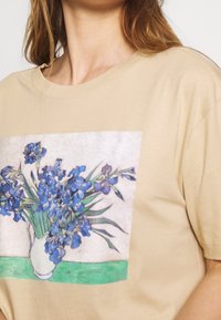 Beige t-shirt med ett tryckt motiv av blå irisblommor i en vit vas, med gröna detaljer vid basen. Mjuk, avslappnad tyg med en ledig passform.