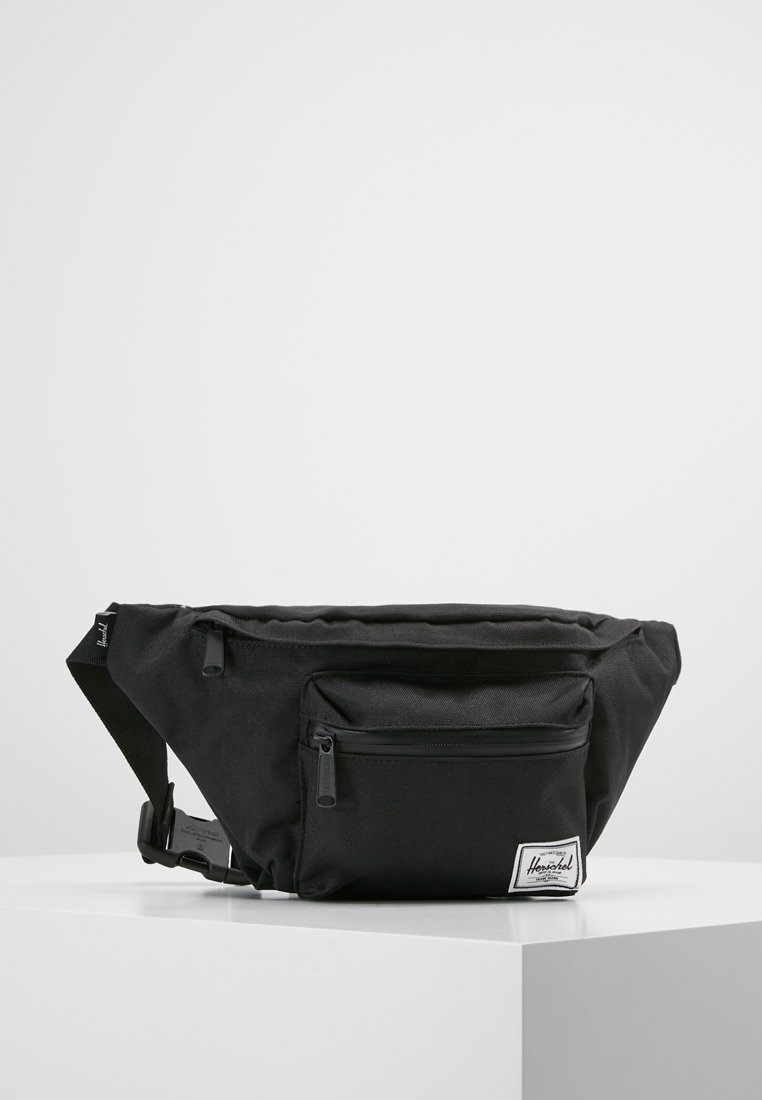 Sac banane en tissu noir avec poche avant zippée, sangle réglable et étiquette blanche de la marque Herschel sur une surface blanche.