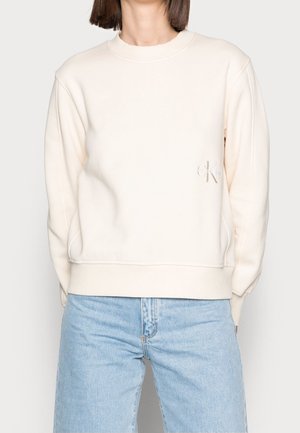 Personne portant un sweat crème Calvin Klein avec un logo discret sur la poitrine et un jean bleu clair, debout devant un fond uni.