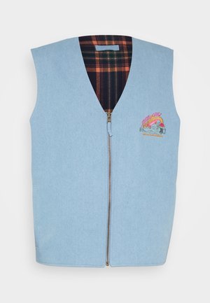 Mand iført lyseblå vest med lynlås over hvid langærmet skjorte, parret med lyseblå jeans og hvide sneakers, stående mod en ensfarvet baggrund.
