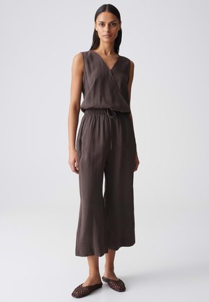 Donna in piedi che indossa un top marrone senza maniche a incrocio, pantaloni larghi abbinati e scarpe marroni intrecciate senza lacci su sfondo semplice.