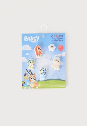 Cinque charm Crocs Jibbitz dei personaggi di Bluey su uno sfondo di cielo azzurro con nuvole, erba e un palloncino rosso.