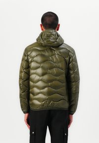 Doudoune vert olive avec capuche, dotée de sections matelassées, d'une texture brillante et de poignets élastiques, portée sur un pantalon sombre.
