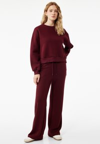 Mujer con cabello rubio ondulado de pie, vestida con una sudadera de manga larga burdeos y pantalones de pierna ancha a juego, con zapatillas beige, sobre un fondo blanco.