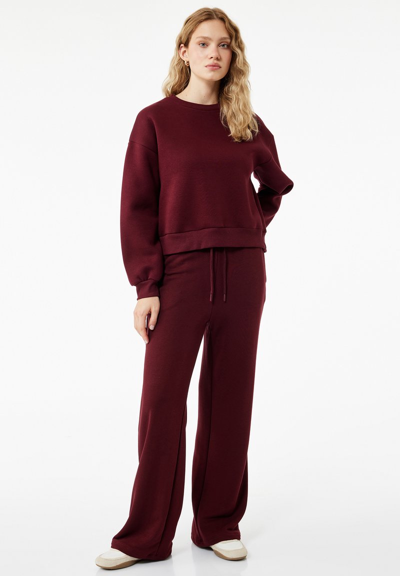 Mujer con cabello rubio ondulado de pie, vestida con una sudadera de manga larga burdeos y pantalones de pierna ancha a juego, con zapatillas beige, sobre un fondo blanco.