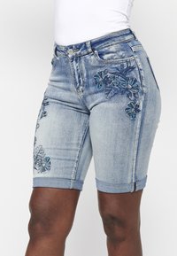 Bermuda en denim bleu clair avec des motifs floraux brodés en teal et marine, présentant des ourlets retroussés et un style à cinq poches.