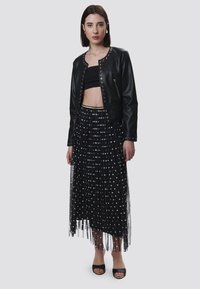 Veste en cuir noire avec des détails cloutés, associée à une jupe noire à pois blancs et à franges, portée avec des talons ouverts noirs.