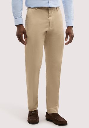 Mężczyzna w beżowych spodniach, jasnoniebieskiej koszuli eleganckiej i ciemnobrązowych mokasynach stoi z rękami swobodnie opuszczonymi wzdłuż ciała.