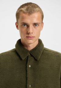 Olivgrüne Fleecejacke mit Kragen, Druckknopfverschluss und strukturiertem Oberflächenmaterial. Das Design beinhaltet ein weiches, warmes Material.