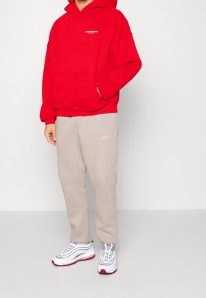 Red hooded sweatshirt with front pocket, paired with beige sweatpants featuring a pocket. Footwear includes white and red sneakers with mesh accents.