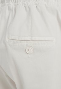 Pantaloni bianchi in cotone con una tasca posteriore con chiusura a bottone e dettagli di cucitura sottili. Vita elastica nella parte superiore.