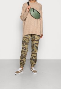 Person in beiger, oversized Strickpullover, mit camouflaged Jogginghose, braunen Sneakers und hält den grünen Umhängetaschenriemen nah am Brustbereich.