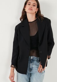 Černý tailored blejzr s klopovým límcem, zapínáním na jeden knoflík a manžetami s květinovým vzorem, kombinovaný s průsvitným topem a džíny.