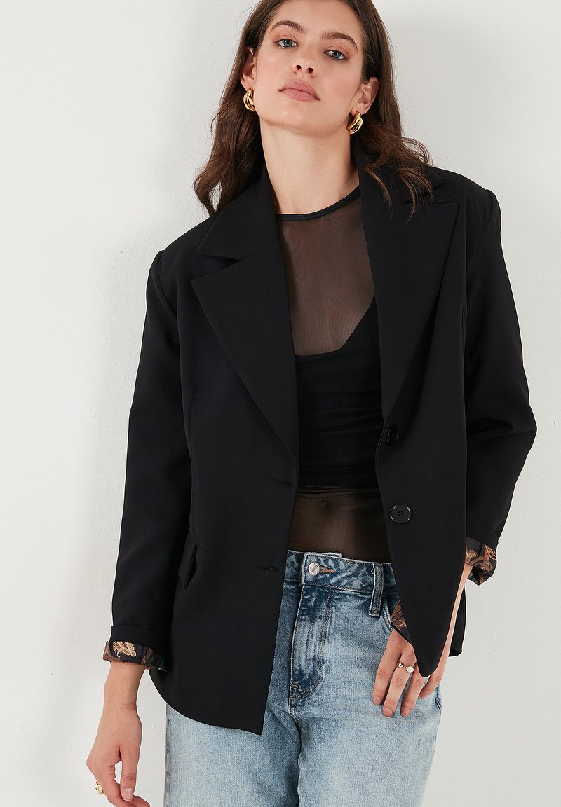 Černý tailored blejzr s klopovým límcem, zapínáním na jeden knoflík a manžetami s květinovým vzorem, kombinovaný s průsvitným topem a džíny.