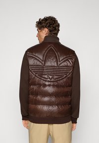 Smeđi podstavljeni prsluk, sjajni materijal, sa velikim izvezenim logotipom na leđima, ispod kojeg se nalazi smeđi pulover i bež pantalone.