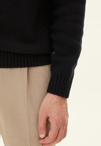 Felpa lavorata a maglia blu navy con dettagli a coste sui polsini, indossata con pantaloni beige, che mettono in mostra il tessuto testurizzato e un primo piano di una mano.