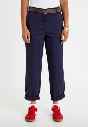 Person wearing cuffed navy pants, white socks, red sneakers, brown belt, and gold bracelets, standing on a white floor against a plain background.