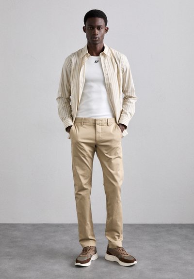 Stribet beige og hvid skjorte med knapper over en hvid top, parret med beige bukser og brune sneakers med teksturerede detaljer.