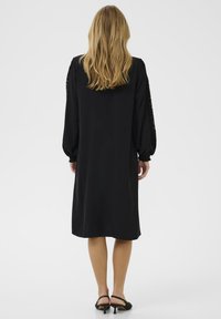 Vestido negro con mangas largas, tejido texturizado y un ajuste relajado. Detalles de encaje en las mangas, de largo justo por encima de la rodilla y con un acabado suave.