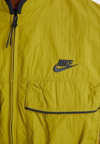 Strålande gul Nike-jacka med dragkedja fram, svart broderad logotyp på bröstet och en ficka med lock på vänster sida.