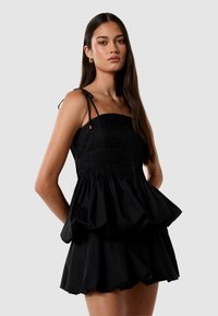 Robe peplum noire avec un corsage smocké, des fines bretelles et un ourlet froncé. Fabriquée dans un tissu lisse, avec des accents en matériel doré.