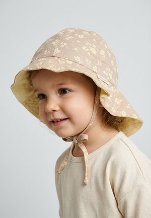 Toddler wearing a beige floral sunhat with chin ties and a cream-colored shirt, smiling slightly, against a plain background.