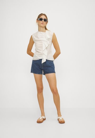 Top blanco sin mangas con un frente con volantes, combinado con shorts de denim azul y sandalias crema con detalle de nudo. La modelo lleva gafas de sol grises.