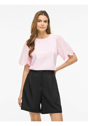 Femme portant une blouse rose pâle avec des manches en dentelle festonnée et un short noir taille haute et ajusté, debout avec une main sur la hanche.