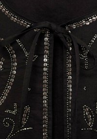 Zwarte stoffen kleding met ingewikkelde kralenwerk langs de randen, met cirkelvormige en geometrische patronen, vastgemaakt met een strik aan de voorkant.