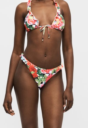 Vrouw draagt een bikini met bloemenpatroon, gerimpelde randen en een voorstrik op het bovenstuk, voor een effen, lichte achtergrond.